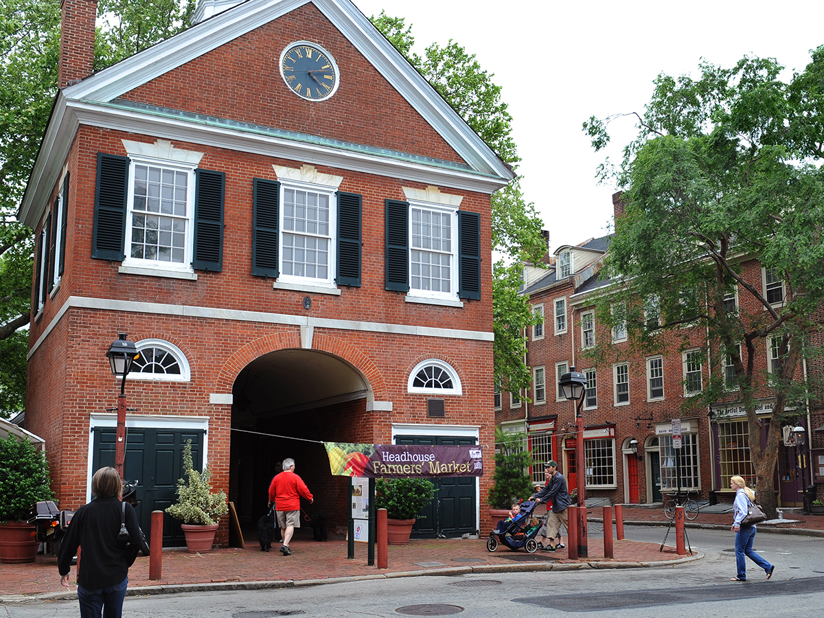 People walk through the street to enter a brick building with 3 windows and a clock on the exterior. The building hosts the Headhouse Farmers' Market.