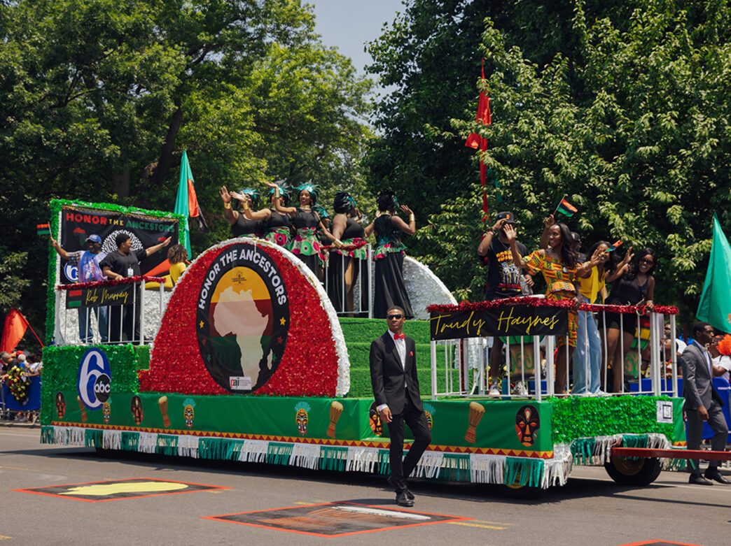 Des personnes sur un char saluent la foule lors de la Juneteenth Parade à Philadelphie. Le char est décoré de vert et de rouge avec l'inscription "Honor the Ancestors" sur le côté.