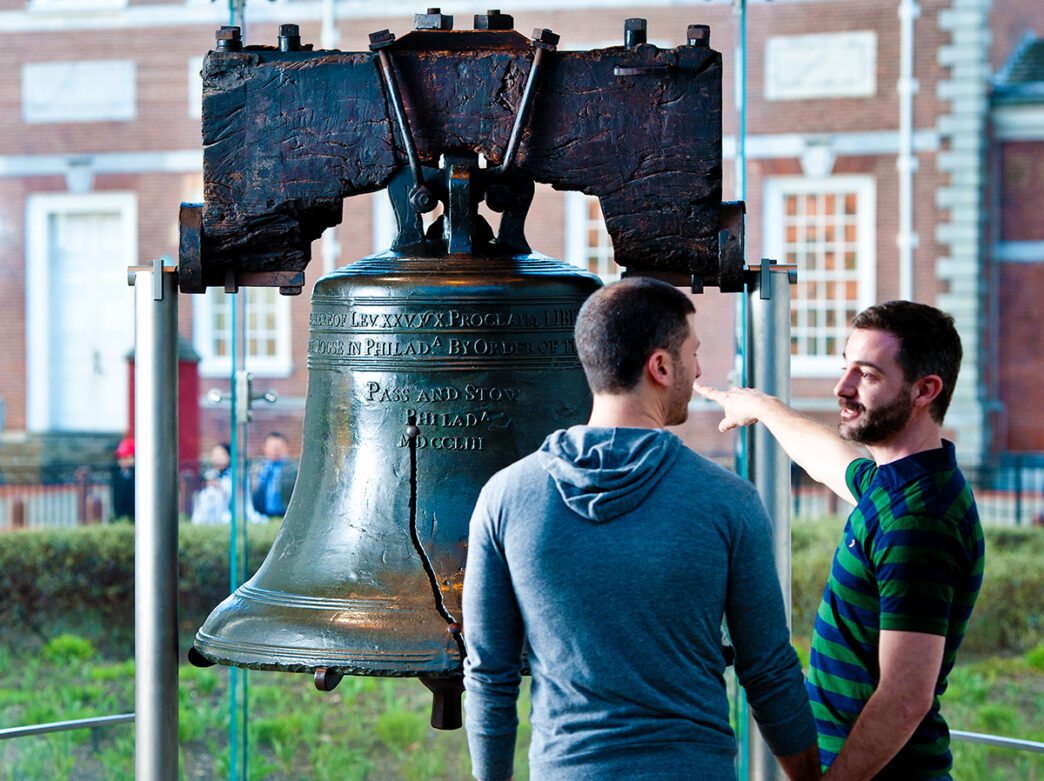 A couple holding hands stand facing the Liberty Bell while one person points to the bell.