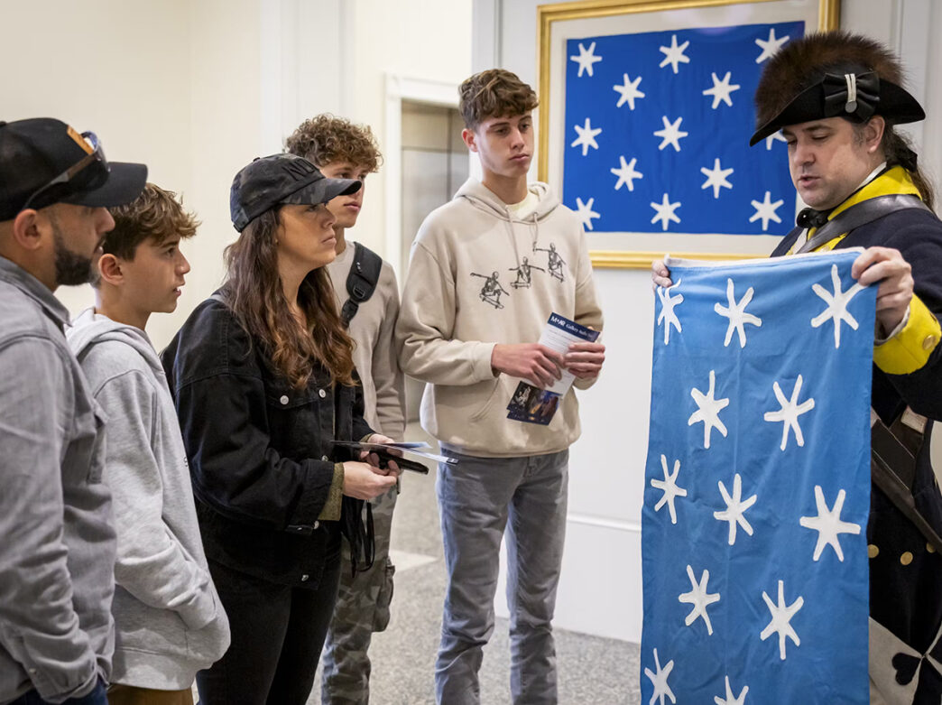 A Revolutionary War impersonator holds up a replica of Washington's Headquarters Flag in front of a family visiting the Museum of the American Revolution in Philadelphia.