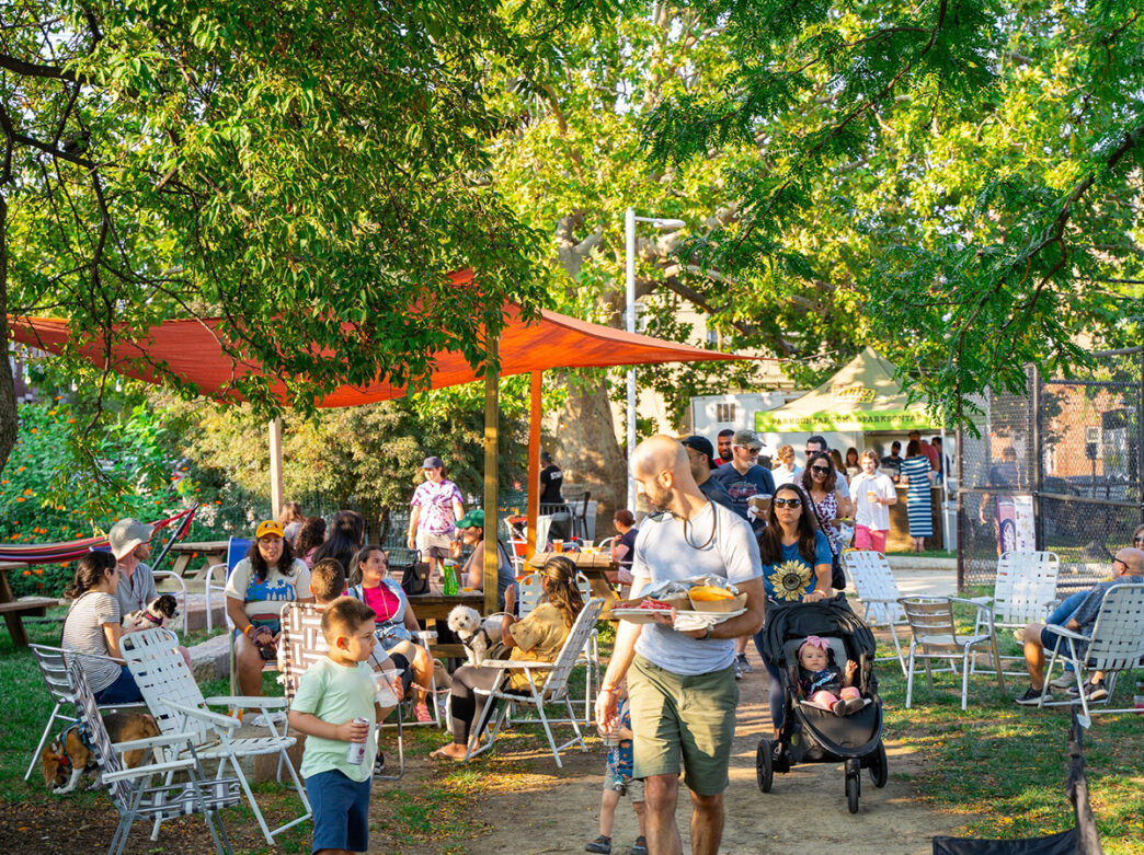 People attend Parks on Tap at Columbus Square Park in Philadelphia. People sit in lawn chairs and walk around with food and drinks.