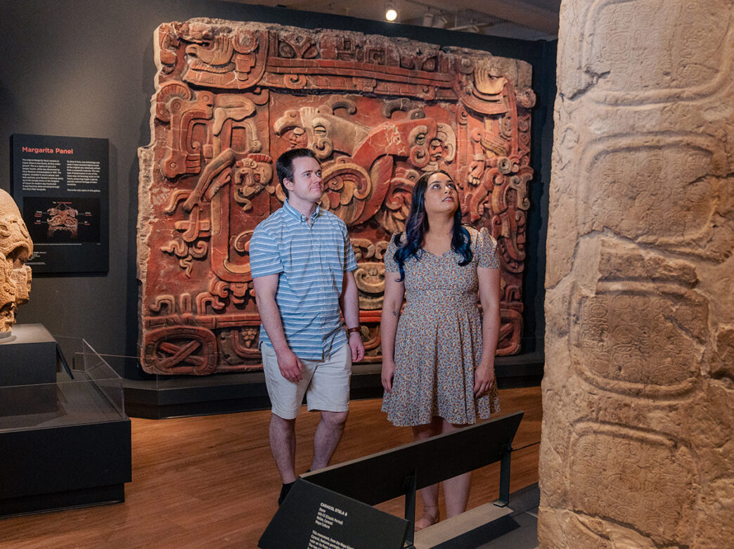 Two museum visitors explore the Mexico and Central America Gallery at the Penn Museum. Various artifacts and sculptures are displayed, including Margarita Panel from the Maya kingdom.