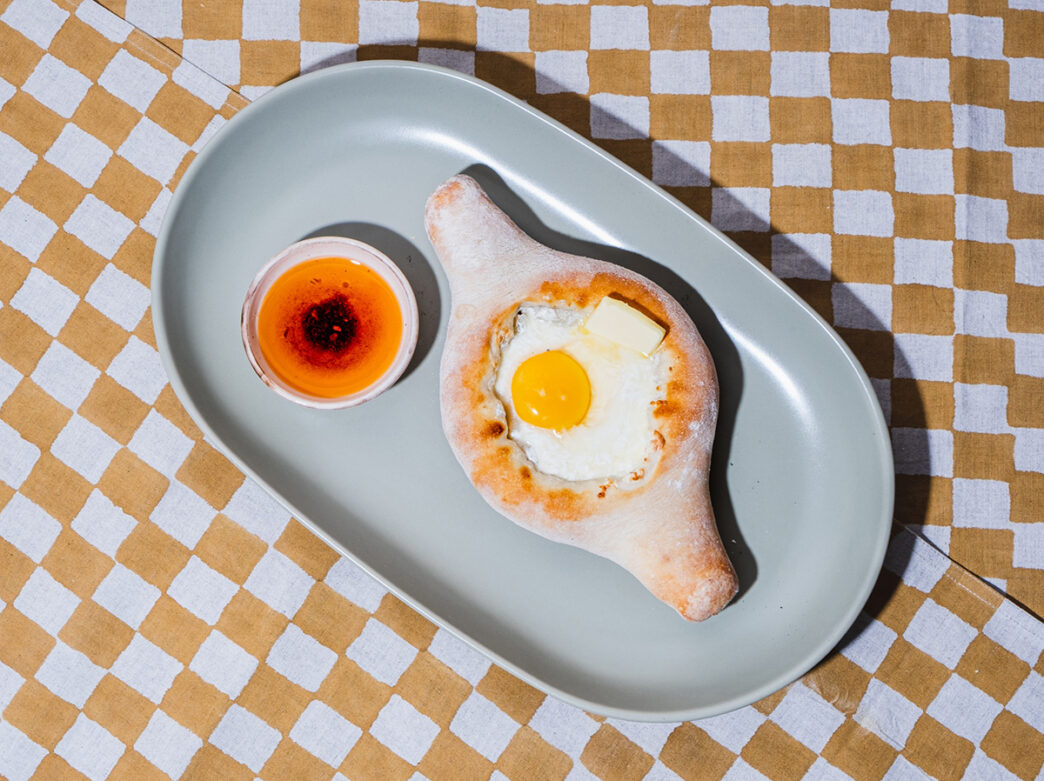 A traditional khachapuri on a gray plate is displayed on a yellow and white checkered tablecloth.