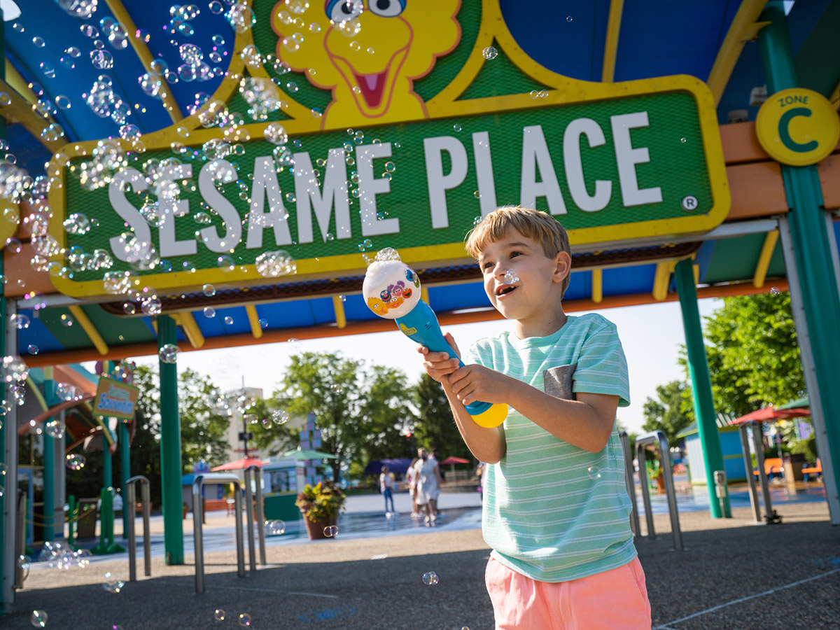 A young person plays with a bubble wand in front of the Sesame Place front gate.