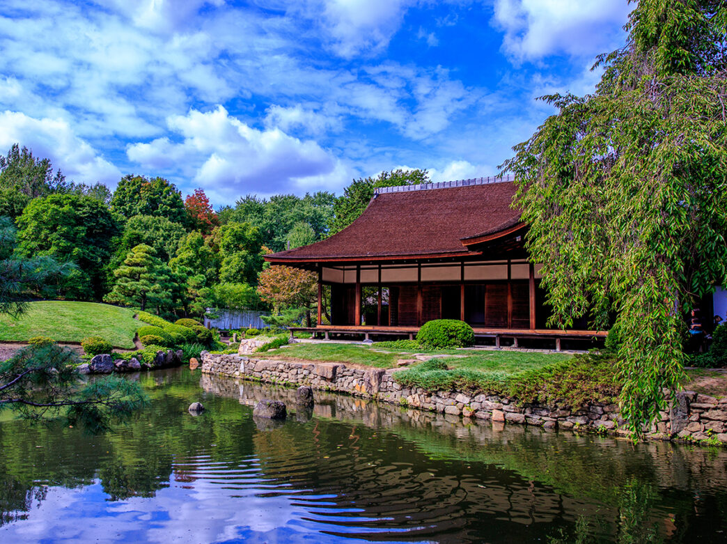 An image of a house, pond and garden at the Shofuso Japanese Cultural Center surrounded by green trees and grass.