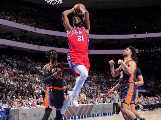 Joel Embiid van de 76ers springt met een basketbal naar de hoepel terwijl drie spelers van de Knicks toekijken.