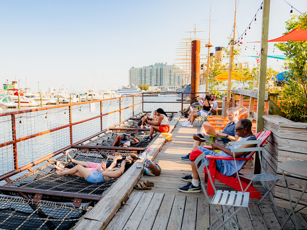 People sit in Adirondack chairs on a wooden deck and lay in netted hammocks over the Delaware River at Spruce Street Harbor Park.