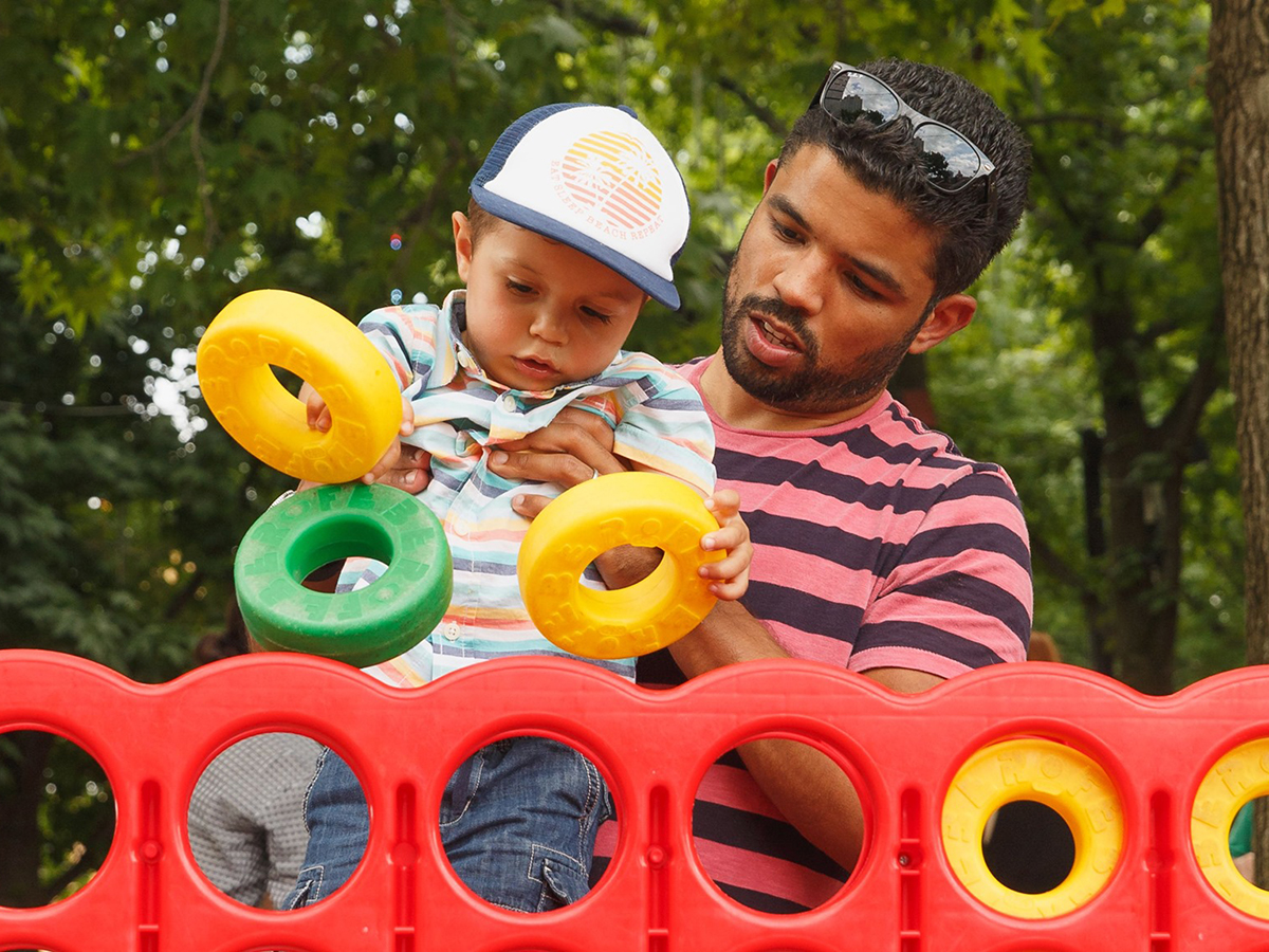 A person holds a child while the child puts giant Connect Four pieces into the game at Spruce Street Harbor Park.