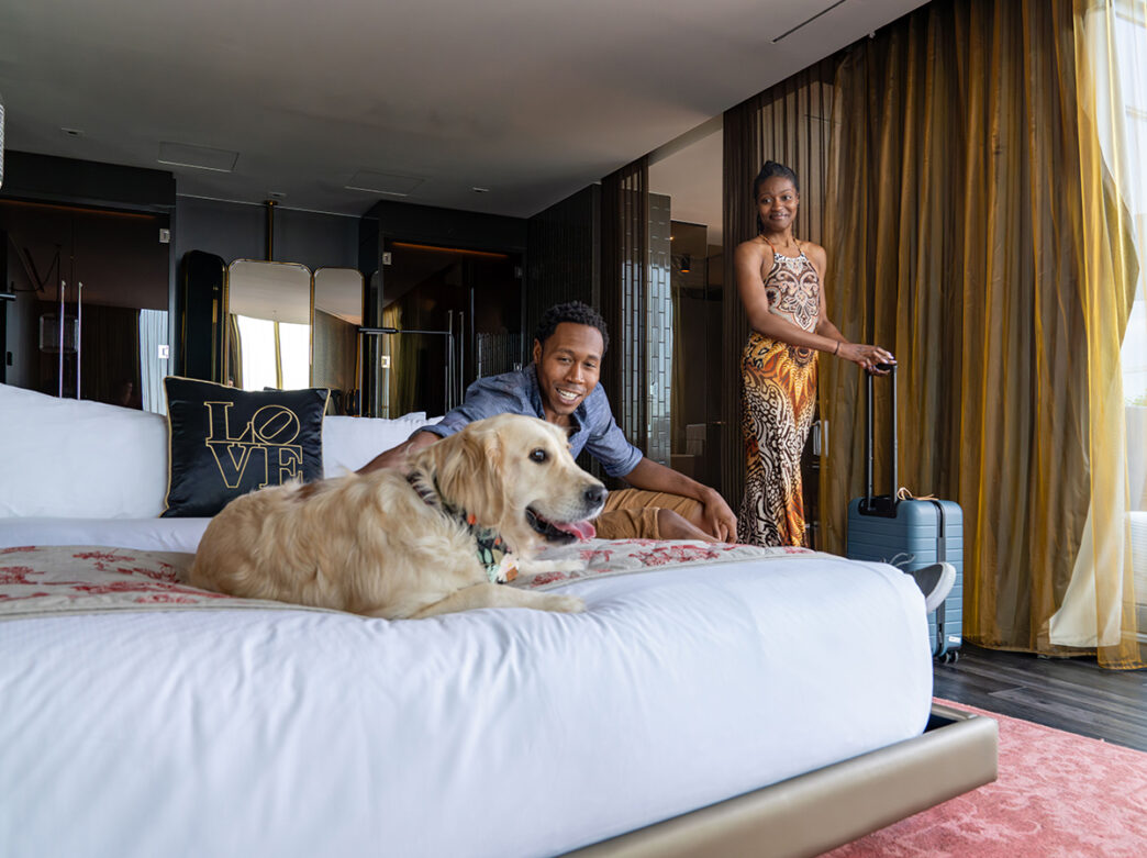 Two overnight guests roll their suitcases into a guest room while their dog lays down on the bed at The W in Philadelphia.