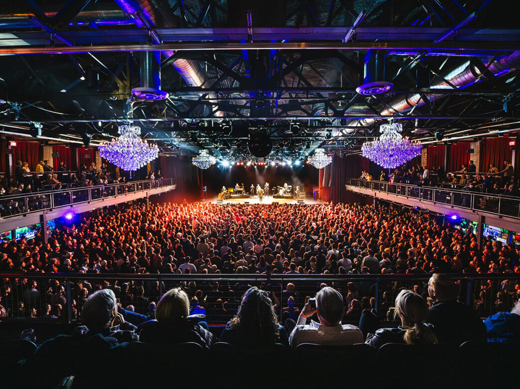 A view of the concert stage at The Fillmore from the rear balcony overlooking the crowd on main floor with glass chandeliers hanging from the ceiling.