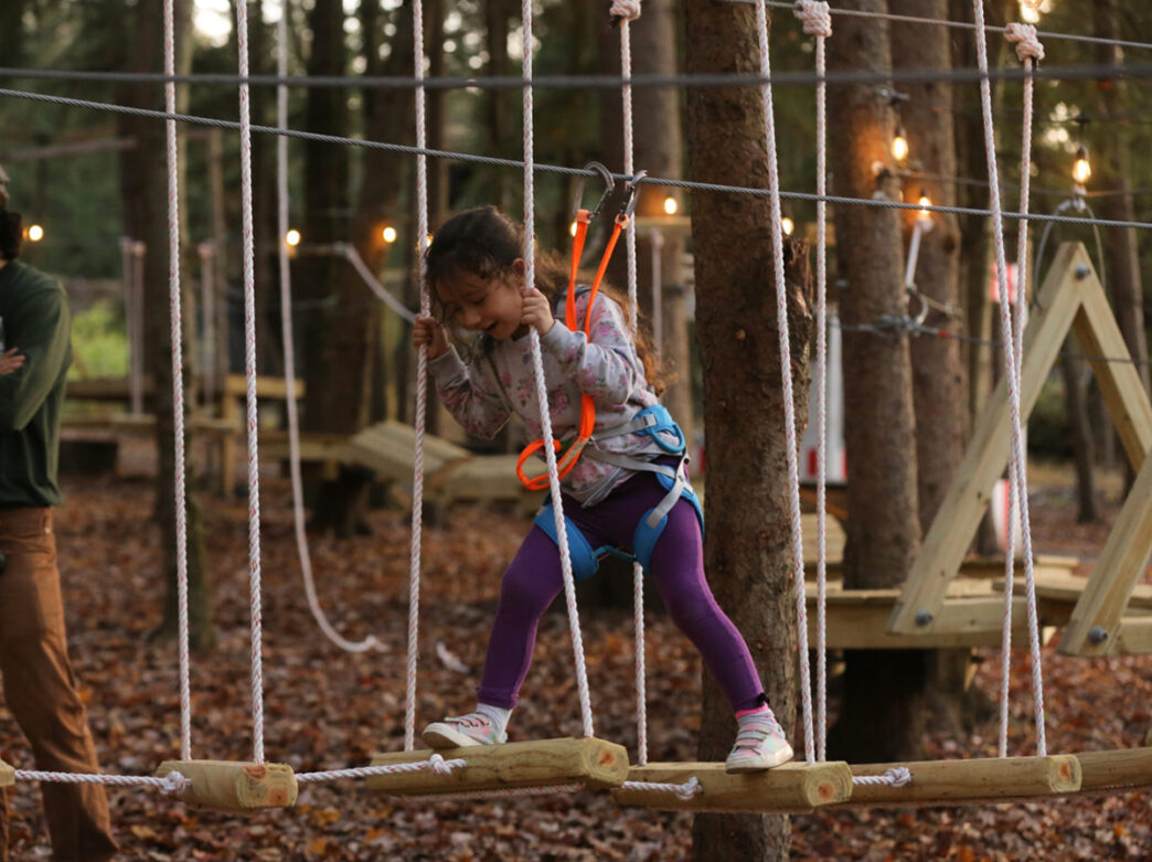 A child wearing a harness walks across wood planks suspended above the ground.