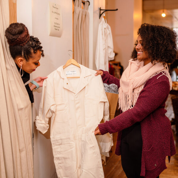 A young woman wearing a burgundy sweater and pink scarf hands her friend a tan jumpsuit to try on in Old City's Vagabond.