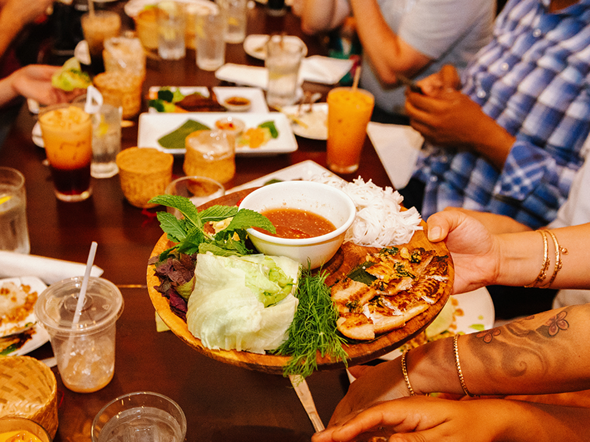 A hand holds out a plate of Thai food over a table of drinks and plates while people dine at Vientiane Cafe in Philadelphia.