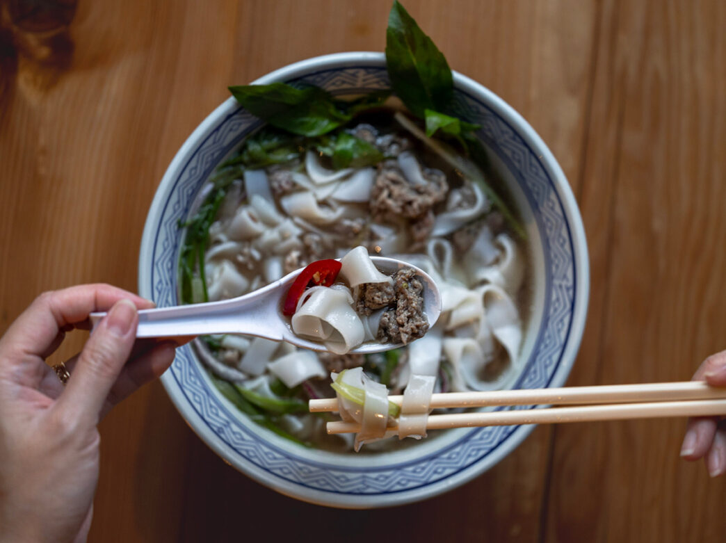 A person holds a pair of chopsticks and a spoon over a bowl of beef pho at Bánh Mì and Bottles in Philadelphia.