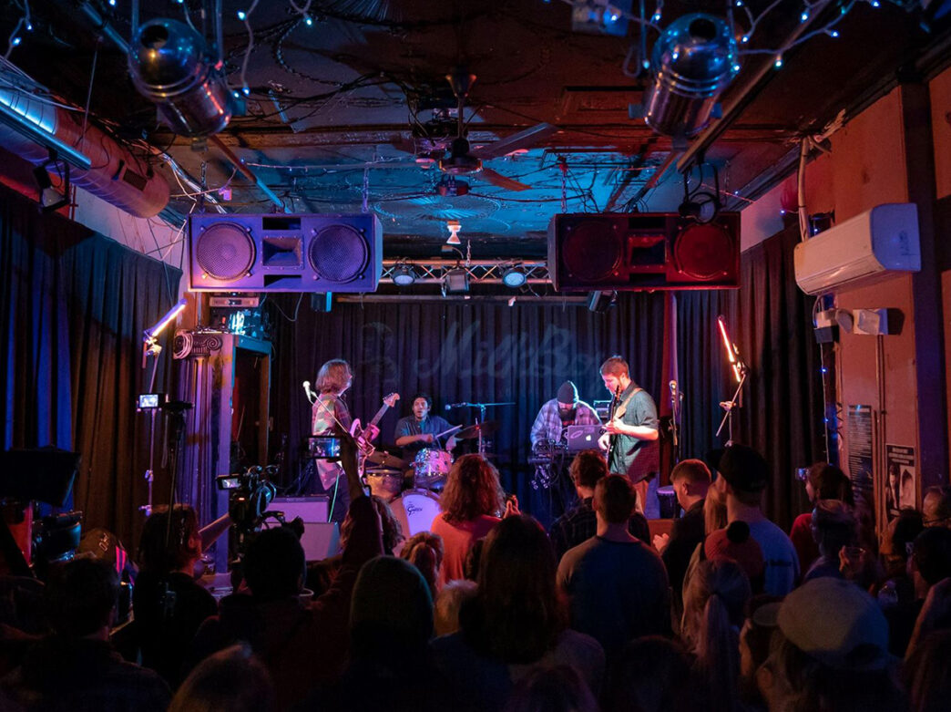A crowd faces the stage while a band performs at MilkBoy in Philadelphia. Large speakers hang from the ceiling.