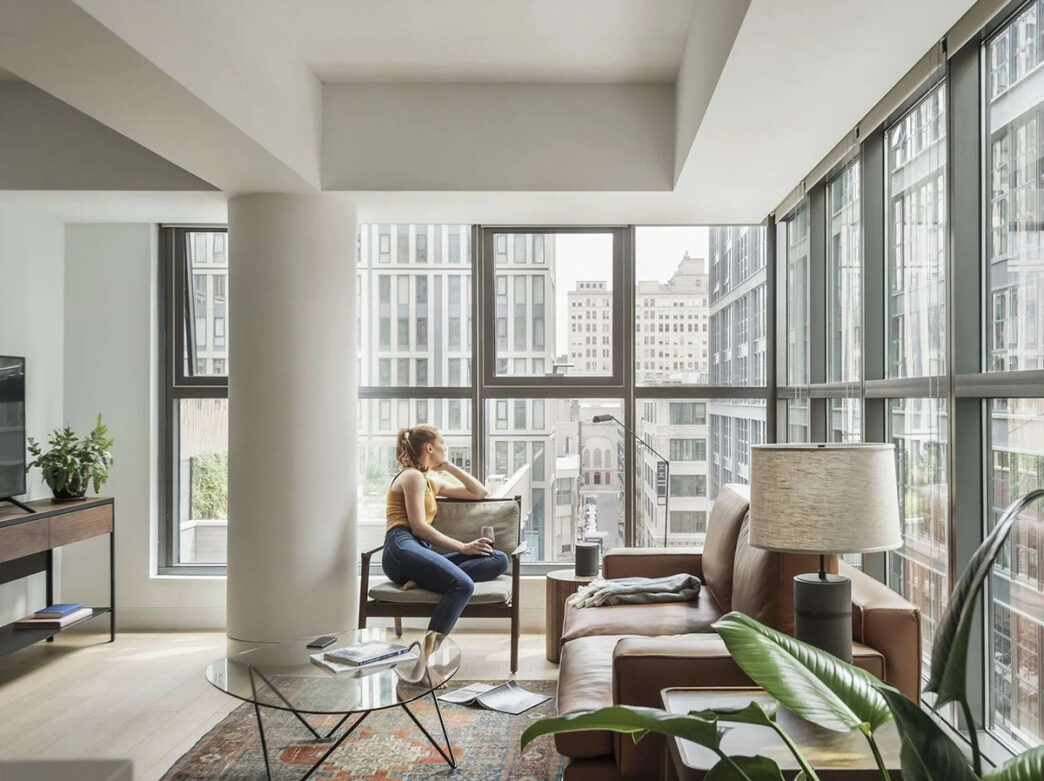 A women sits in the living room inside an apartment at ROOST East Market while drinking a glass of wine and looking out the windows.