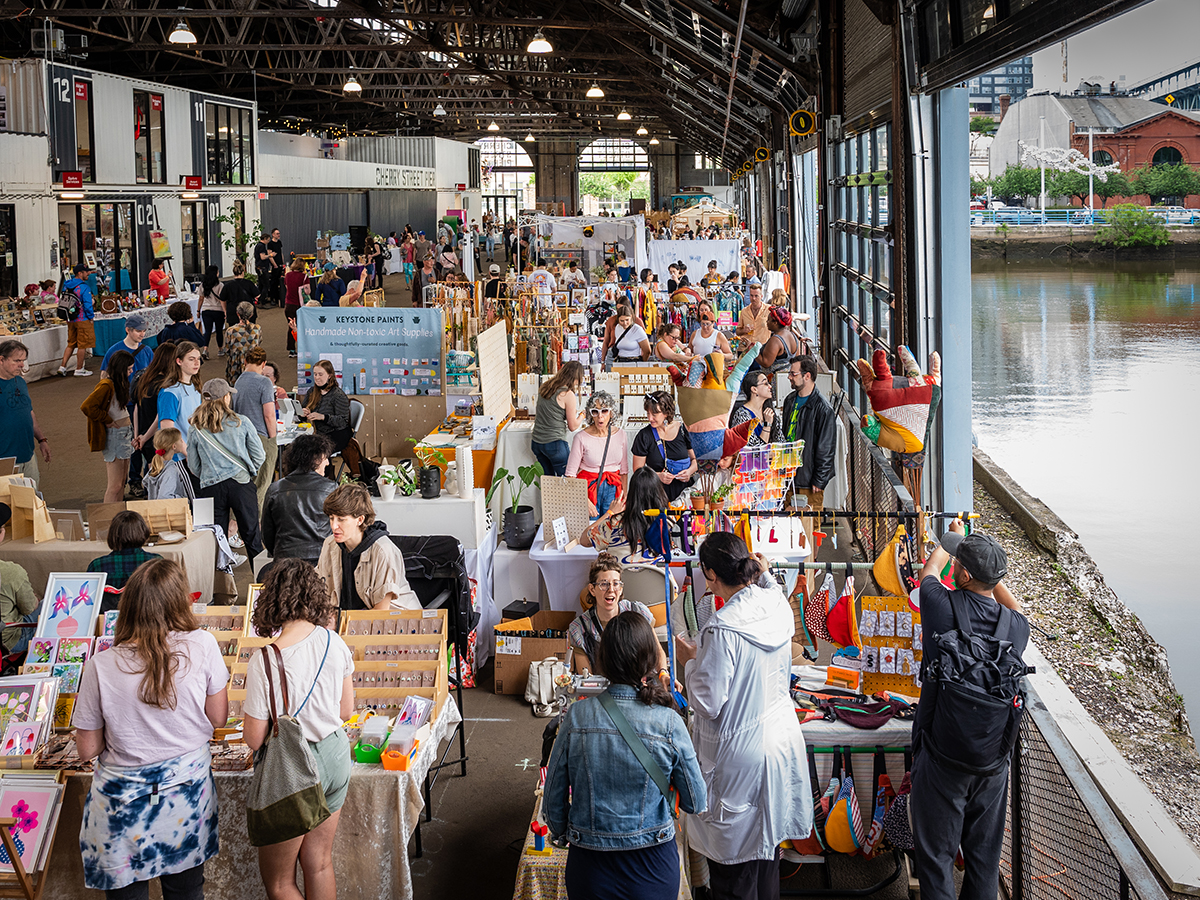 People shop at the vendor booths at Art Star Craft Bazaar at Cherry Street Pier in Philadelphia. The garage doors of the market are open and overlook the Delaware River.