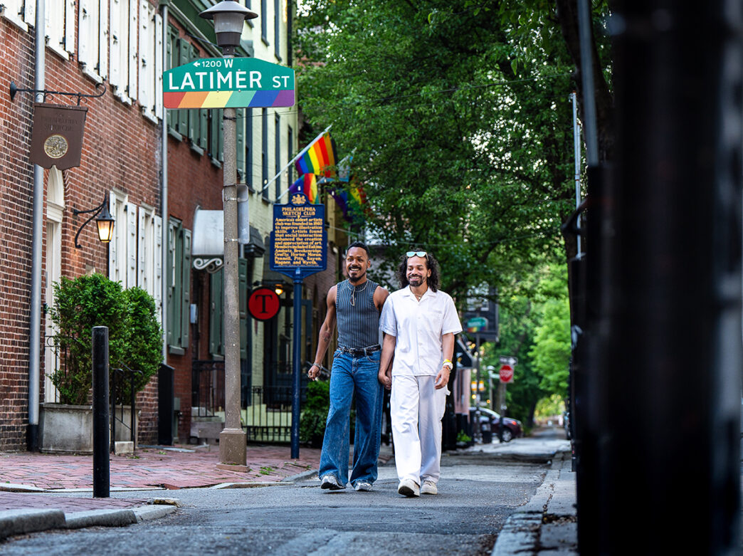 Two people walk down the street holding hands with rainbow flags in the background.
