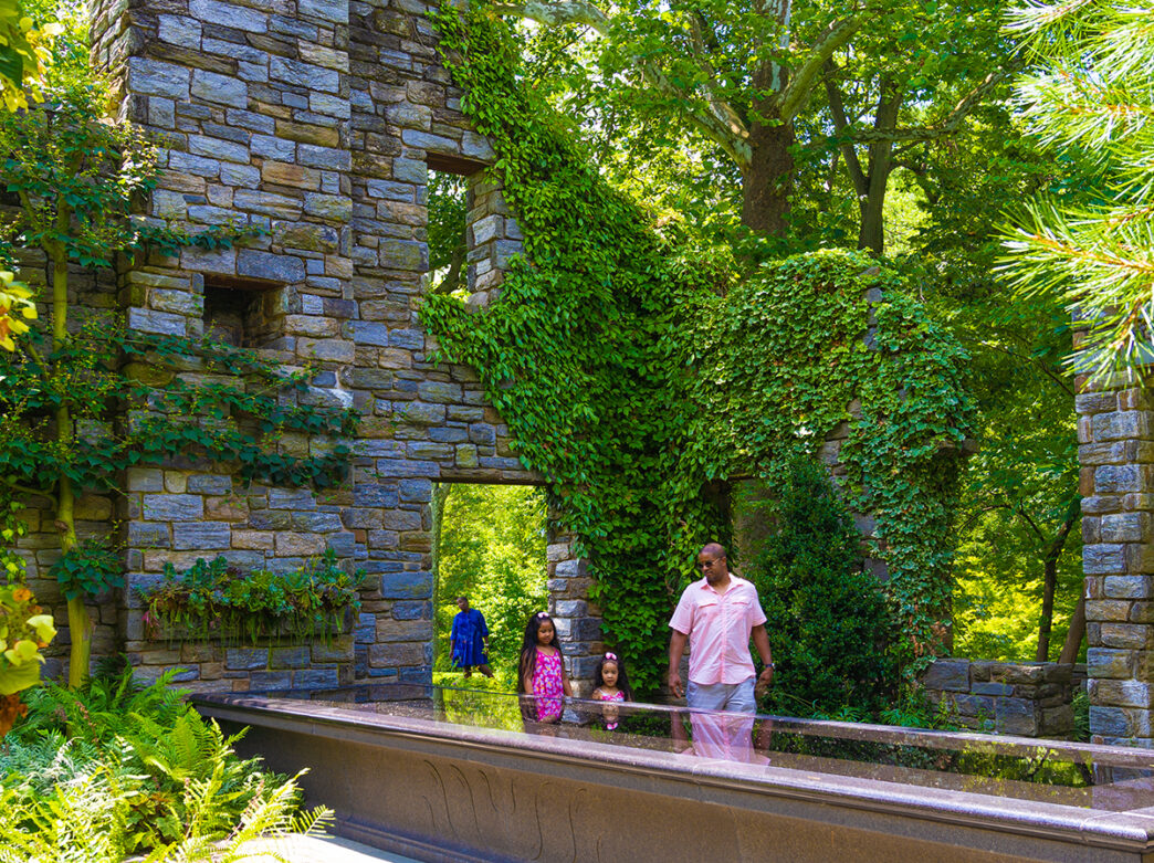 A parent and two children walk through the Ruin Garden at Chanticleer. The stone ruins are surrounded by tall trees and covered in ivy.