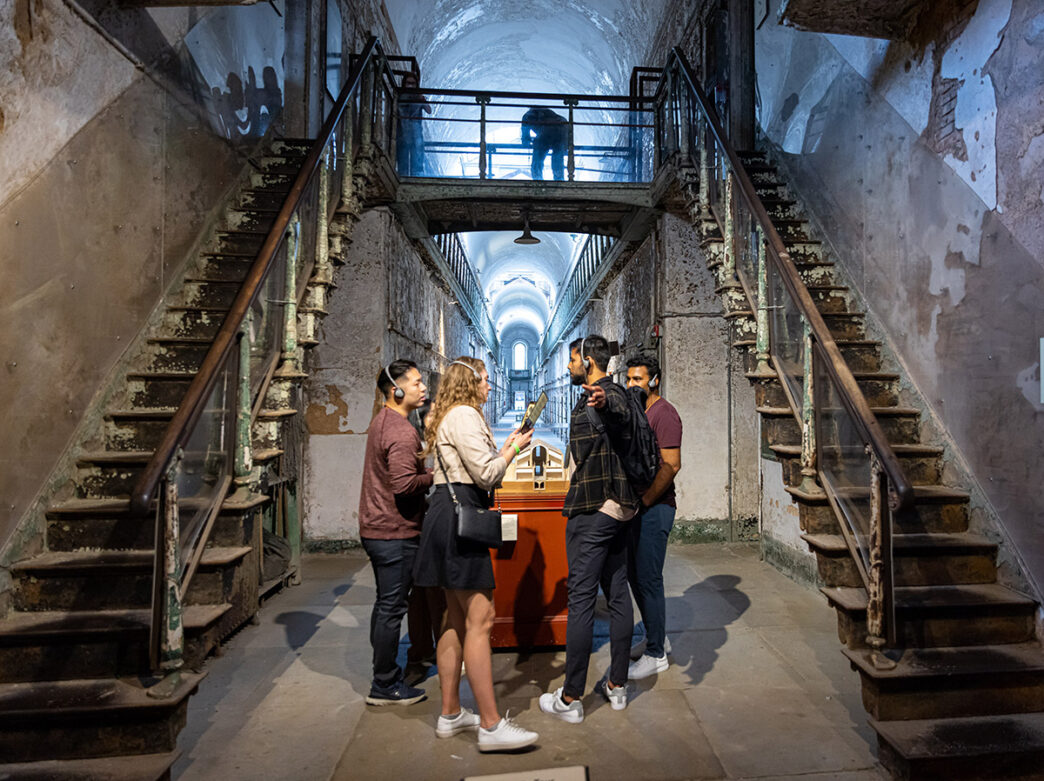 A group of four young, casually dressed Eastern State Penitentiary visitors listen to an audio tour as they explore the institution's halls.