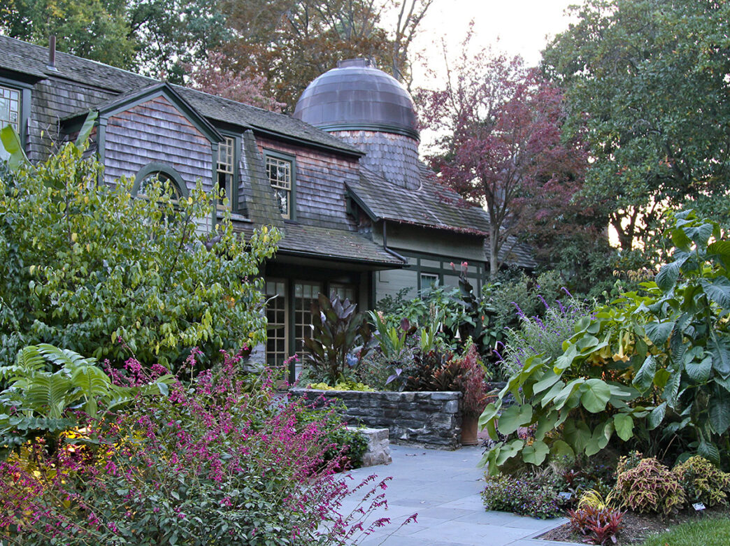 A slate walkway through a garden of plants and shrubs leads up to a gray building with cedar shake siding.
