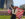 A person stands facing the camera holding an American flag behind their back. In the background is Philadelphia's Independence Hall.