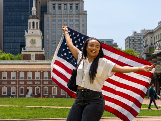 A person stands facing the camera holding an American flag behind their back. In the background is Philadelphia's Independence Hall.
