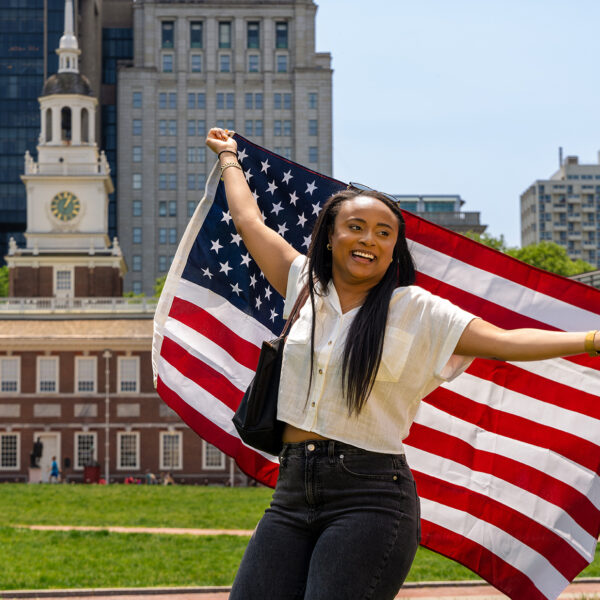 Une personne se tient face à la caméra, tenant un drapeau américain derrière son dos. En arrière-plan, on aperçoit le Independence Hall de Philadelphie.