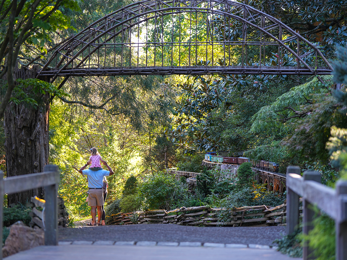 A father carries a child on their shoulders as they walk past the Garden Railway model trail at Morris Arboretum in Philadelphia.