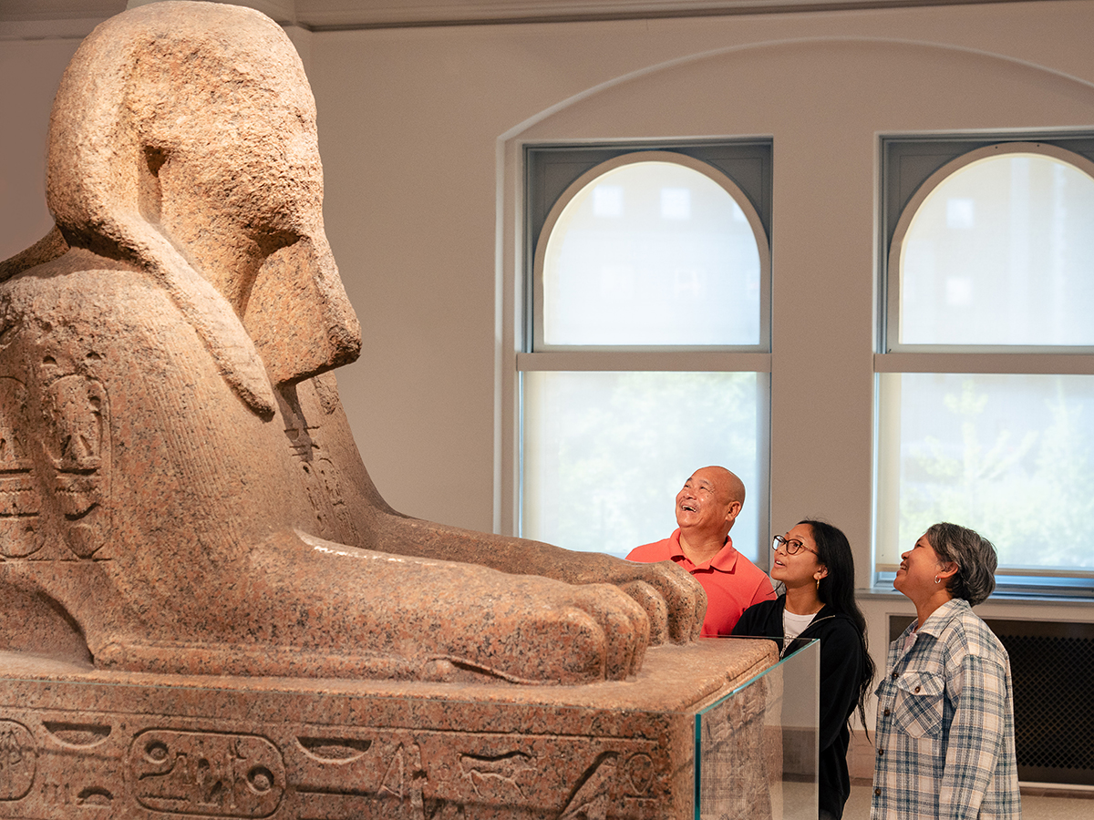 A family of three smile and look up towards a tan granite sphinx at the Penn Museum in Philadelphia.