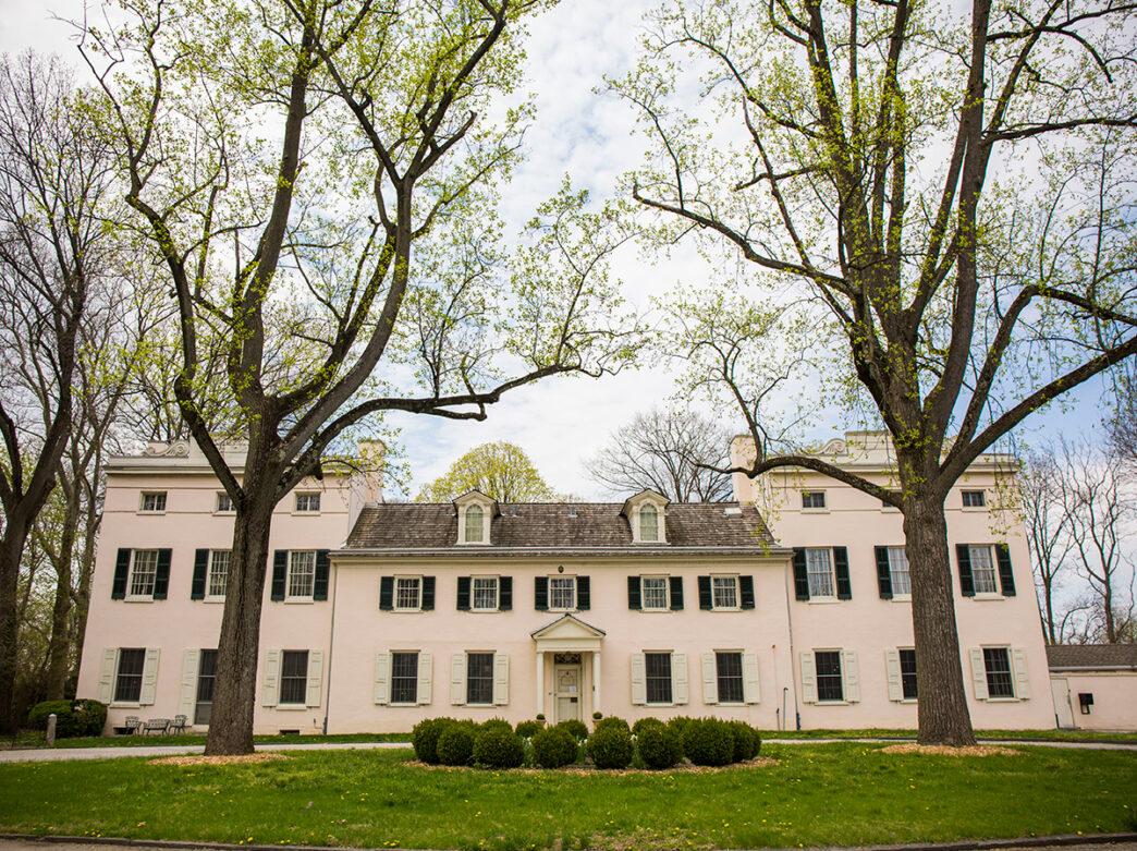 A light pink mansion with a green lawn, bushes and large trees starting to bloom.