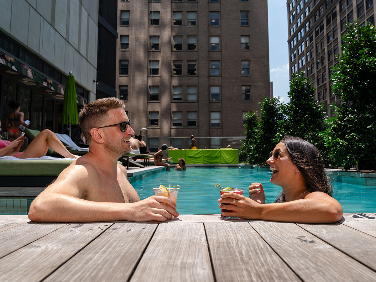 Duas pessoas relaxam na piscina do The W, na Filadélfia. Ambas usam óculos escuros e seguram uma bebida nas mãos.