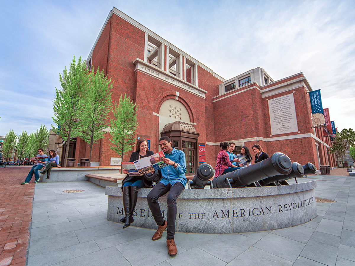 People look at brochures white sitting outside of a large red brick building with replica cannons.