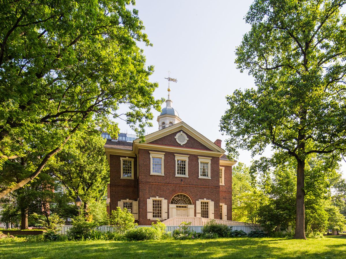 A two story red brick building with a white cupola on the roof and white windows is photographed in between two large green trees.