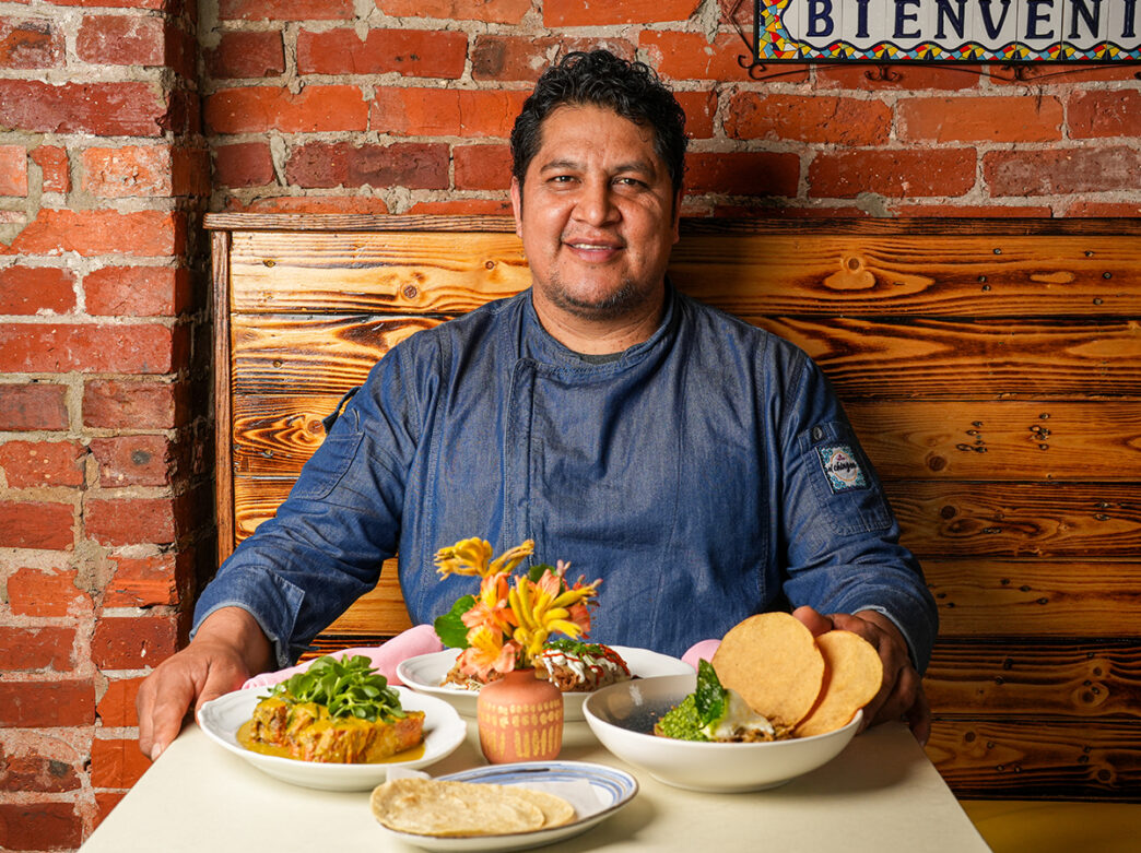 Chef Carlos of El Chingon sits at table and smiles at the camera with several menu items displayed on the table.