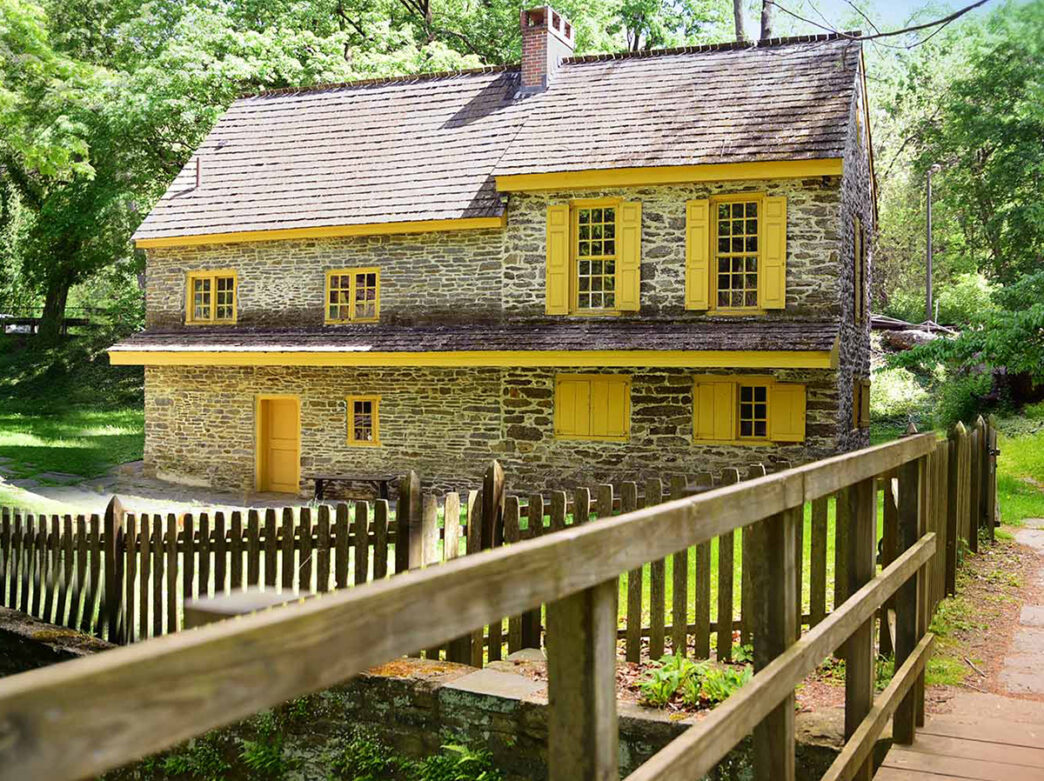 A stone house with yellow windows, shutters and doors sits on a grassy lawn with a wooden fence and walkway.
