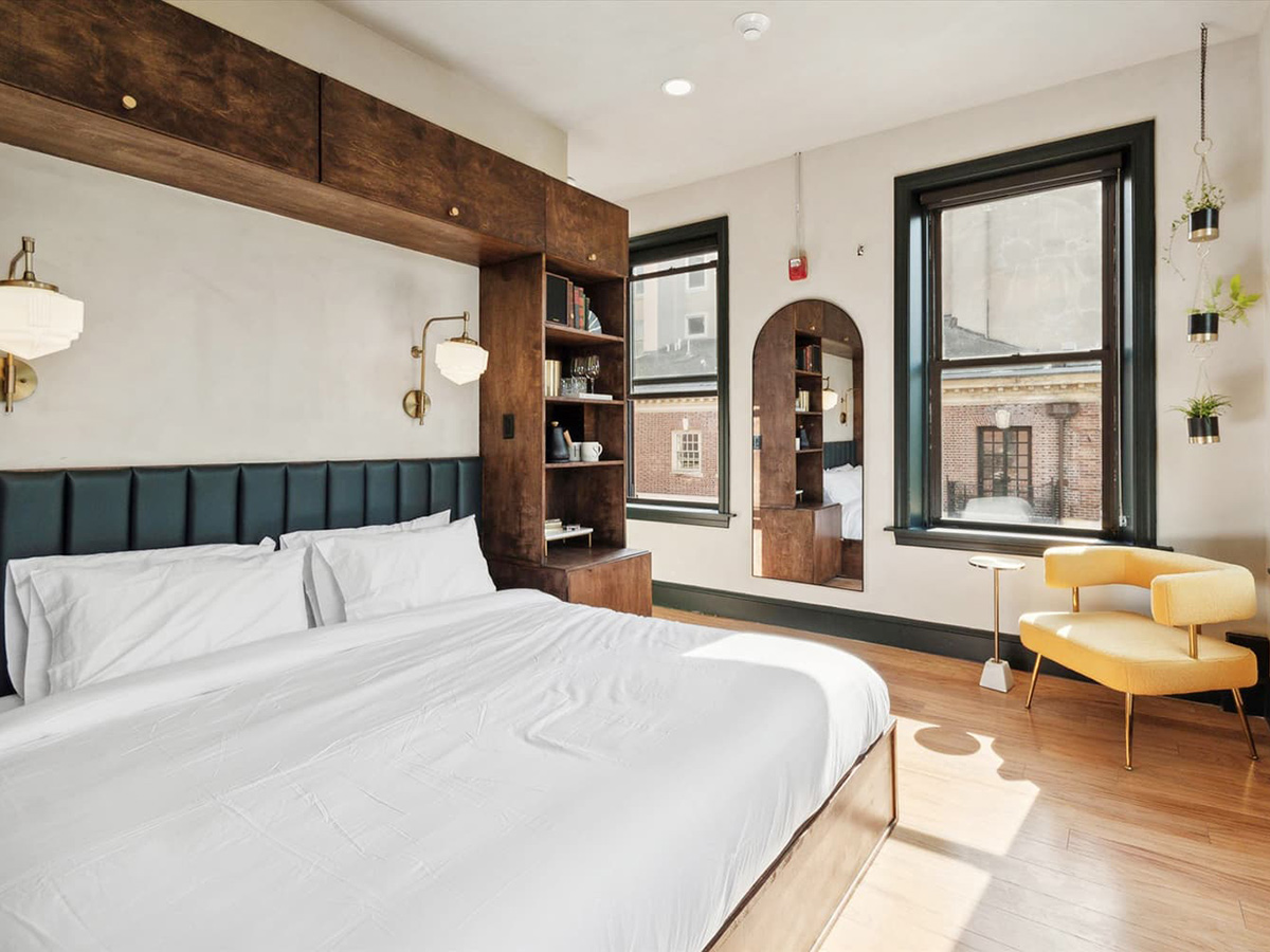 A guest room with a king bed with white linens and decorated in art deco style with large windows and a hanging plant.