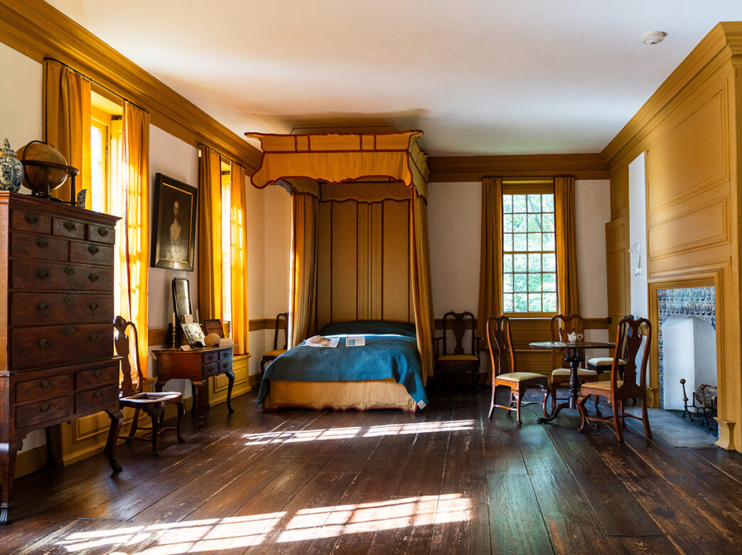 A bedroom with a yellow curtains, molding and accent wall. It is furnished with wood chairs, a dresser and bed with colonial bed hangings.