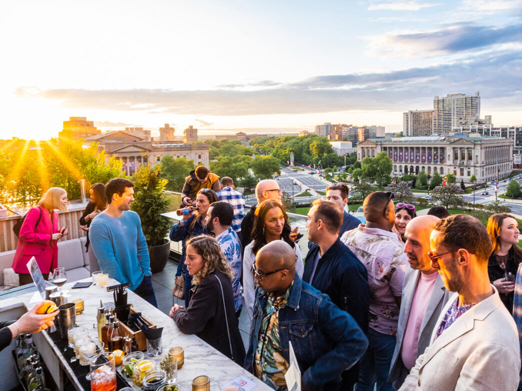 People order drinks at Assembly Rooftop Lounge with a view of the Parkway Central Library in the background as the sun sets.