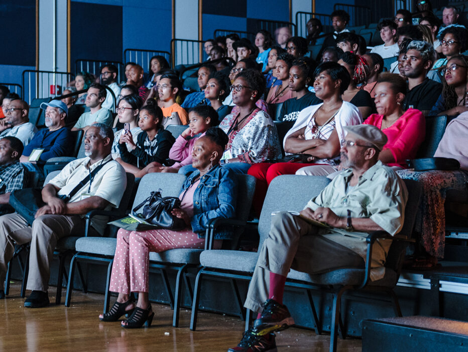 A crowd of people sit in a theater while the lighting from a movie lights up their faces.
