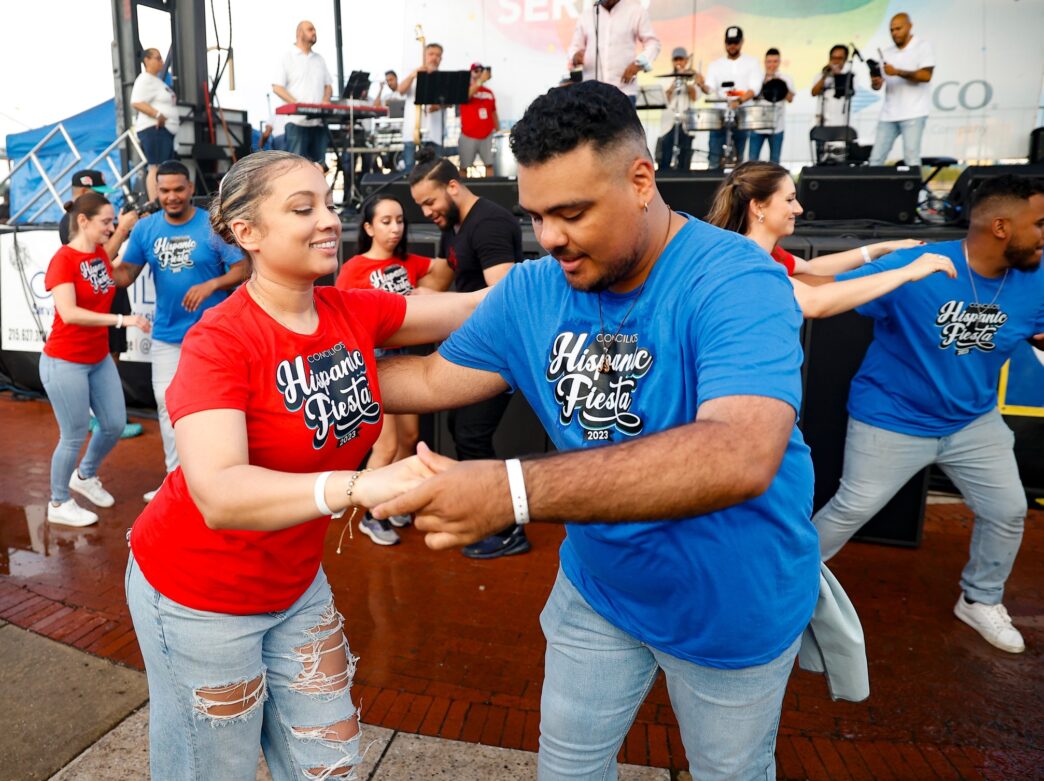 Two people, one wearing a red shirt and one wearing a blue shirt, dance together in front of a band performing on stage.