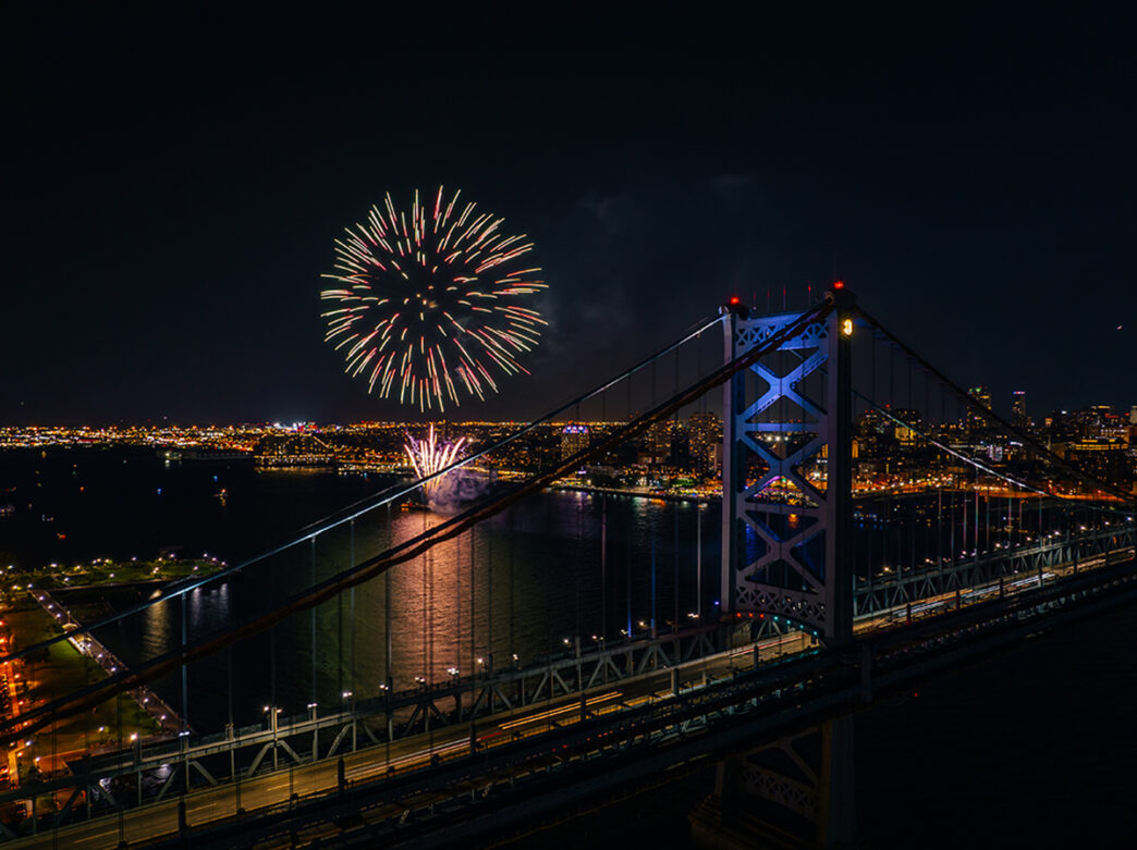 An elevated view over the Delaware River and Benjamin Franklin Bridge at night with a colorful firework exploding in the sky.