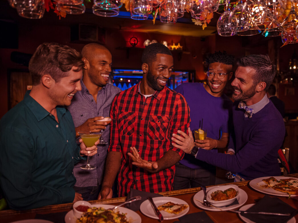 Five people stand at a bar with drinks in their hands with food dishes on the bar top and wine glasses hanging from above.