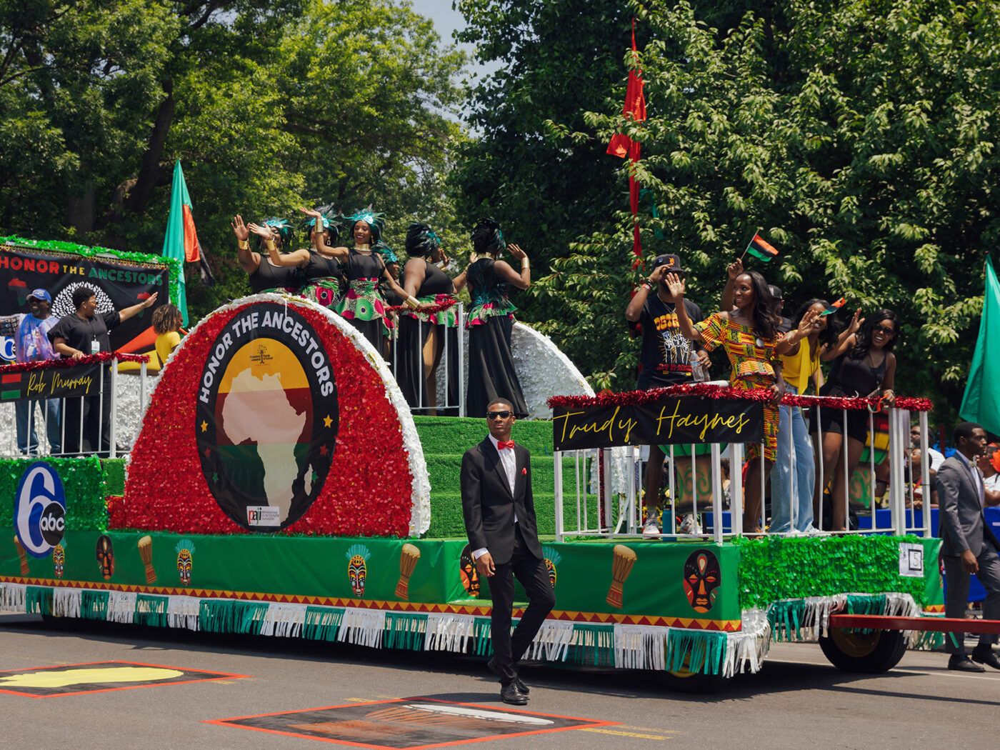 People on a float wave to the crowd during the Juneteenth Parade in Philadelphia. The float is decorated with green and red with "Honor the Ancestors" written on the side.