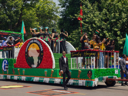 People on a float wave to the crowd during the Juneteenth Parade in Philadelphia. The float is decorated with green and red with "Honor the Ancestors" written on the side.