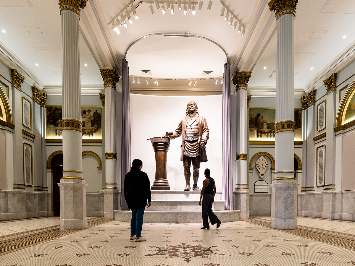 Two people look up at a large bronze statue of Benjamin Franklin displayed in the Masonic Temple in Philadelphia.