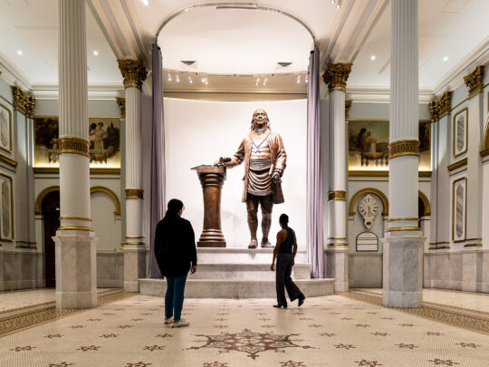 Two people look up at a large bronze statue of Benjamin Franklin displayed in the Masonic Temple in Philadelphia.