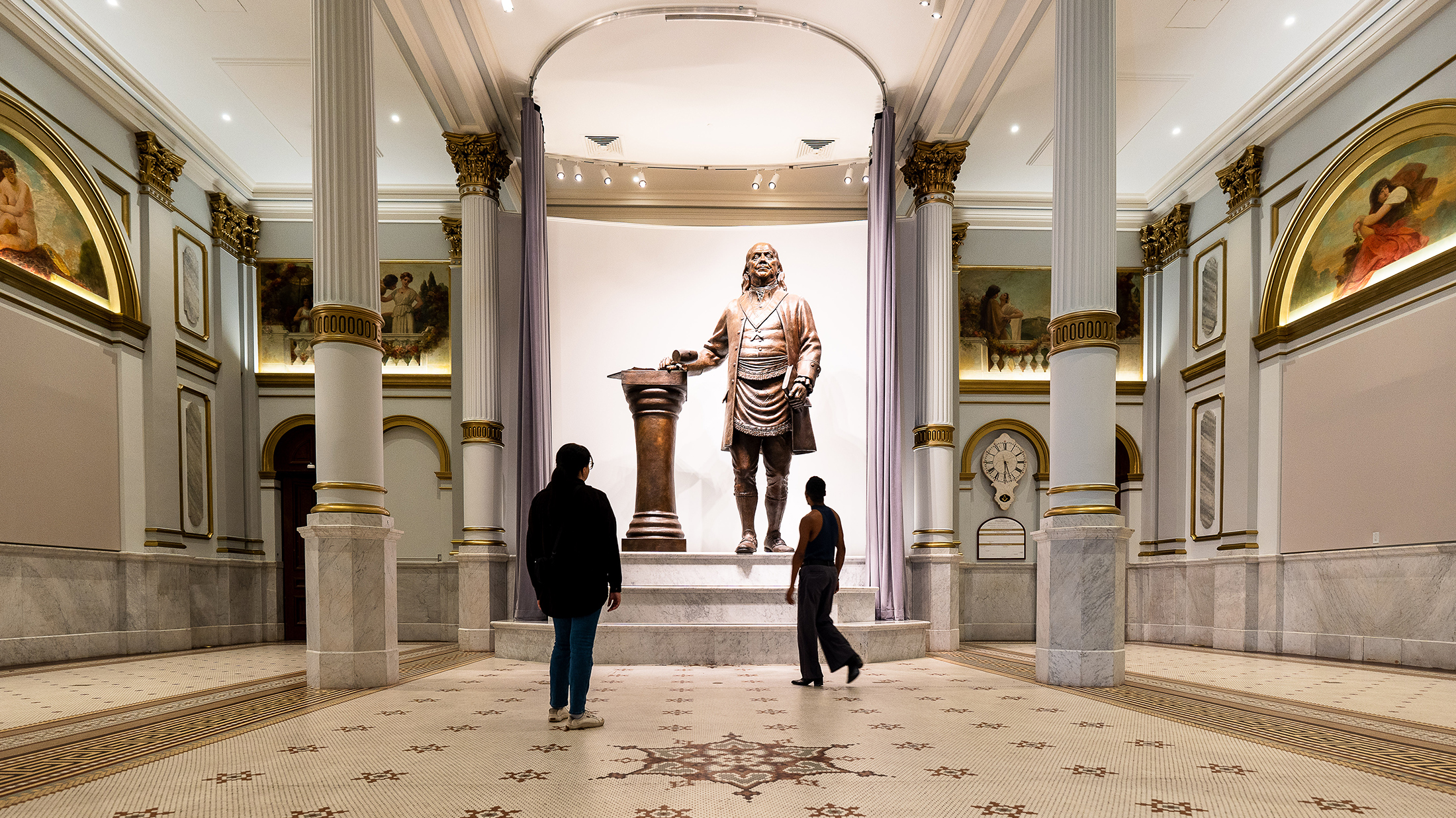 Two people look up at a large bronze statue of Benjamin Franklin displayed in the Masonic Temple in Philadelphia.