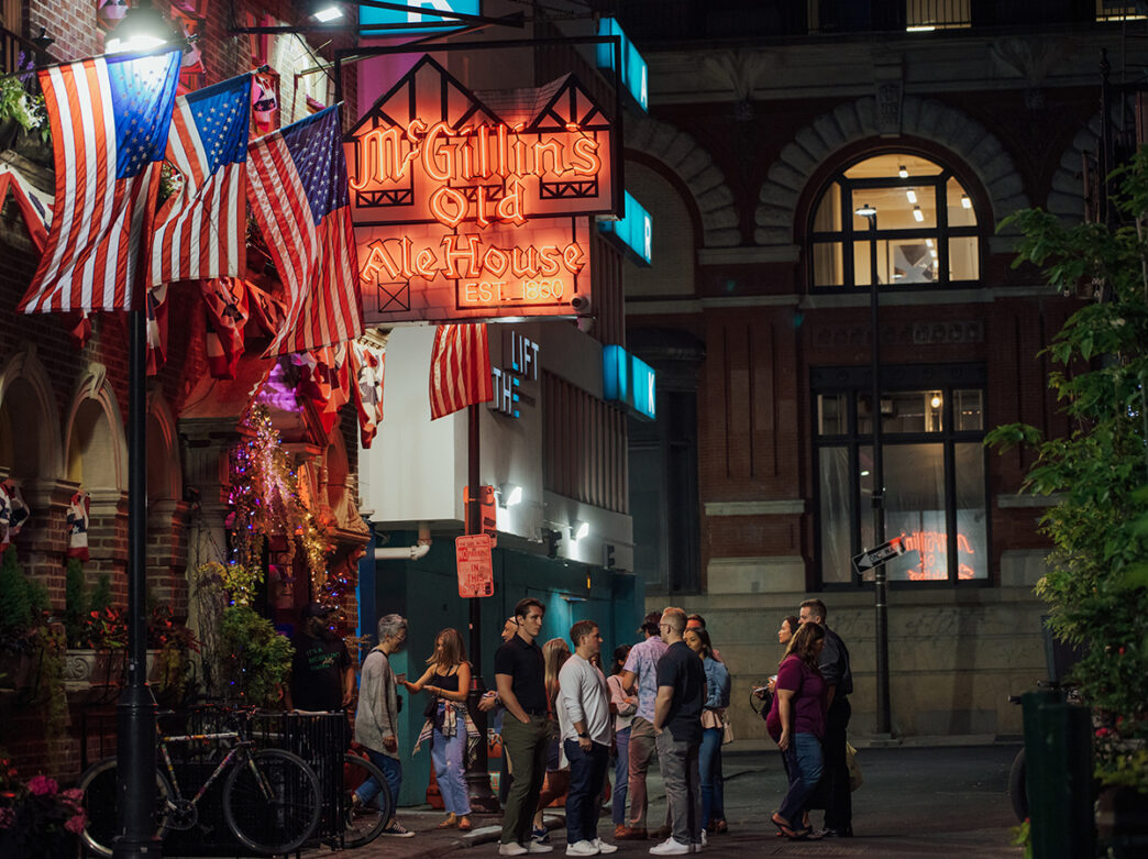 People stand outside and enter McGillin's Old Ale House in Philadelphia. A neon sign and three American flags hang above the bar's entryway.
