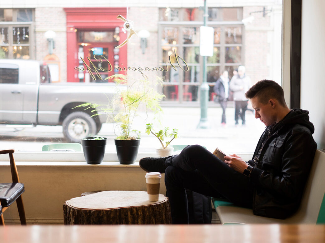 A person sits on a chair in front of a large window while reading a book. A white paper coffee cup sits on coffee table in front of them.